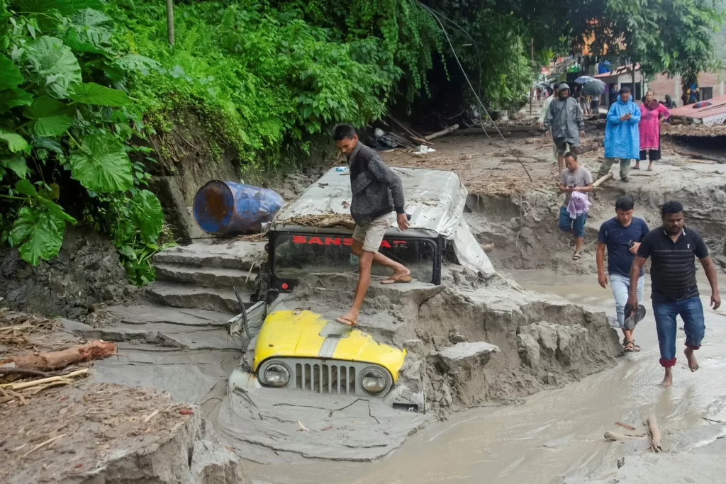 People walk along a street as a jeep is buried in the mud due to the flood at Teesta Bazaar