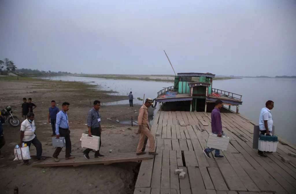 Here’s what you need to know about the world’s largest democratic election kicking off in India 2 Election officers carry Electronic Voting Machines (EVM) on board a ferry to cross the Sowansiri river to reach a polling center on the eve OF elections in Majuli, India, March 26, 2021. (AP Photo/Anupam Nath, File)